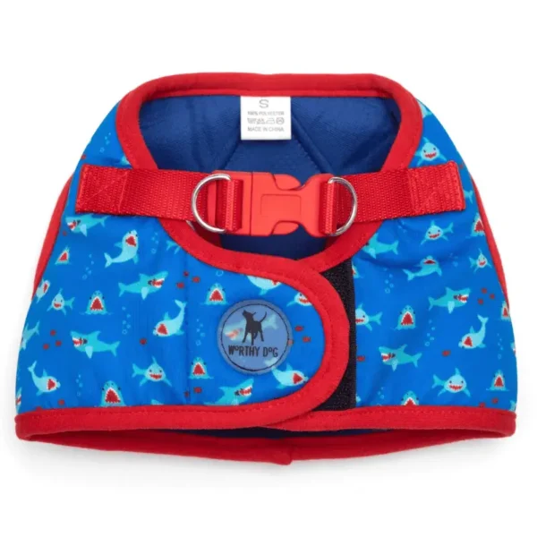 A blue dog harness decorated with shark patterns and a red trim. It features a red buckle and a metal D-ring for leash attachment.