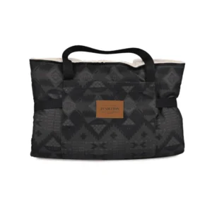Travel dog bed folded up into a black tote bag with a geometric pattern and a leather logo label on the front. It has two handles and a soft, textured lining.