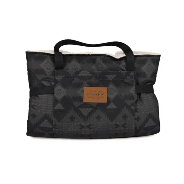 Travel dog bed folded up into a black tote bag with a geometric pattern and a leather logo label on the front. It has two handles and a soft, textured lining.